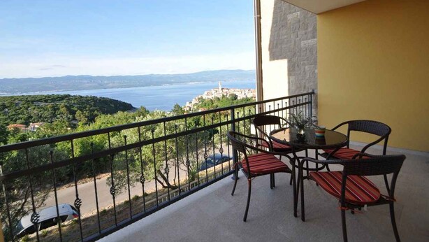 Ein Balkon mit Tisch und Stühlen, der auf eine Küstenlandschaft mit freiem Blick auf das Meer und ferne Hügel blickt.