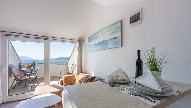Un espace repas moderne avec vue sur la mer, un tableau représentant des bateaux et une bouteille de vin posée sur la table.