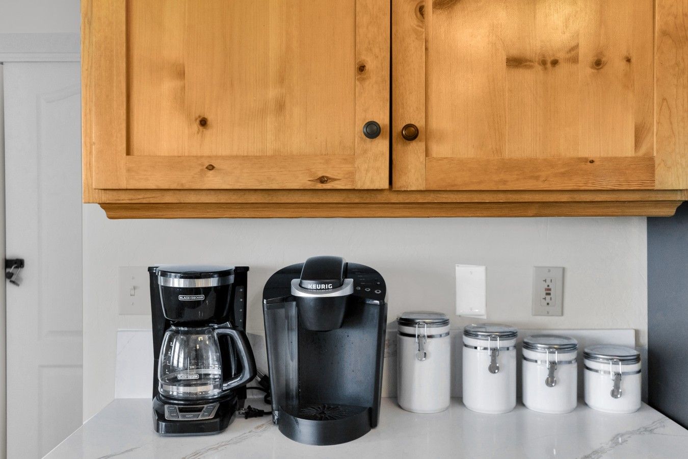 Et køkken med træskabe, en sort kaffemaskine, en Keurig-kaffemaskine og tre hvide beholdere på bordet.