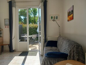 A room with a sofa, a desk, a chair, a dining table, and a sliding glass door leading to an outdoor area with a table and chairs. - "Au Chant des Cigales" – Peaceful Retreat with Large Garden in the Dordogne (Vanxains)