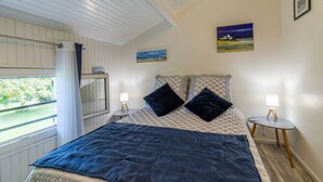 Une chambre à coucher avec un lit, deux tables de chevet avec des lampes, une fenêtre avec des rideaux et deux tableaux accrochés au mur.
