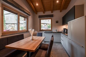 Een moderne keuken met een houten tafel en bank, een koelkast, en twee ramen met uitzicht op een stenen muur en groen.