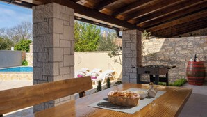 An outdoor patio with a wooden table, stone pillars, a foosball table, and a pool in the background. - Villa with terrace for 2 people (Orbanići)