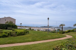 A paved pathway, manicured grass, a lamp post, and a building with a view of the sea and boats.