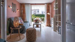 A living room with a sofa, a round ottoman, a table with a vase, and a view of a garden through an open door. - Magnificent T2 Apartment with Sea View and Wi-Fi (Six-Fours-les-Plages)