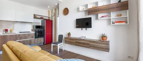Una sala de estar moderna con un sofá, un televisor montado en la pared y una cocina al fondo.