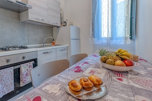 Cozinha com fogão, forno e geladeira. Há uma mesa com uma fruteira e doces.
