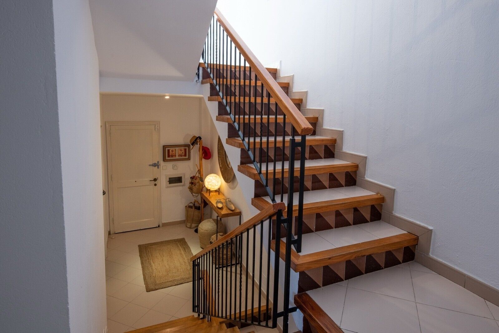 A staircase with wooden steps and black railings leading to a landing with a door, a wall-mounted light, and a small table with a lamp.