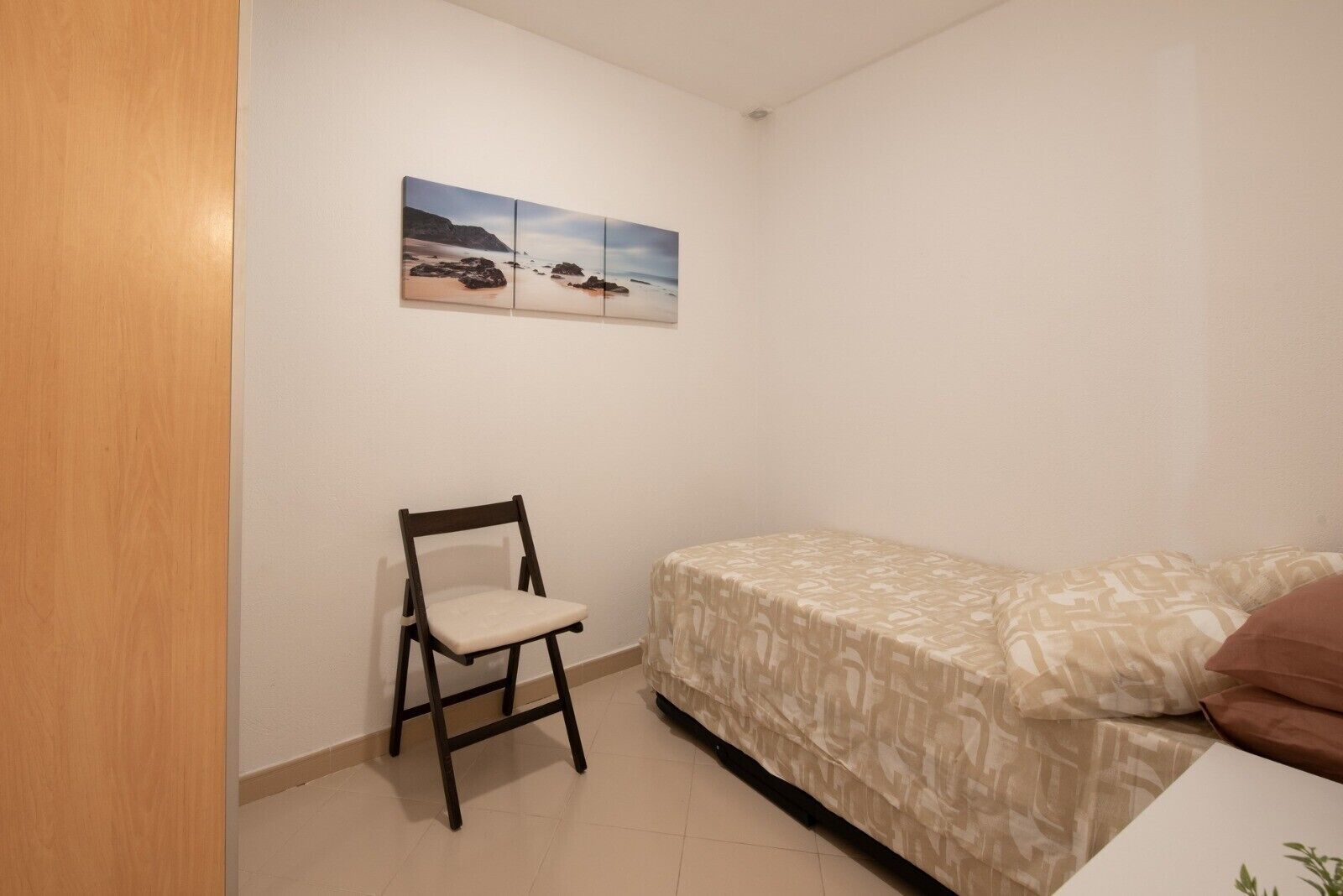 A small, simple room with a bed, a chair, and a wall-mounted picture.