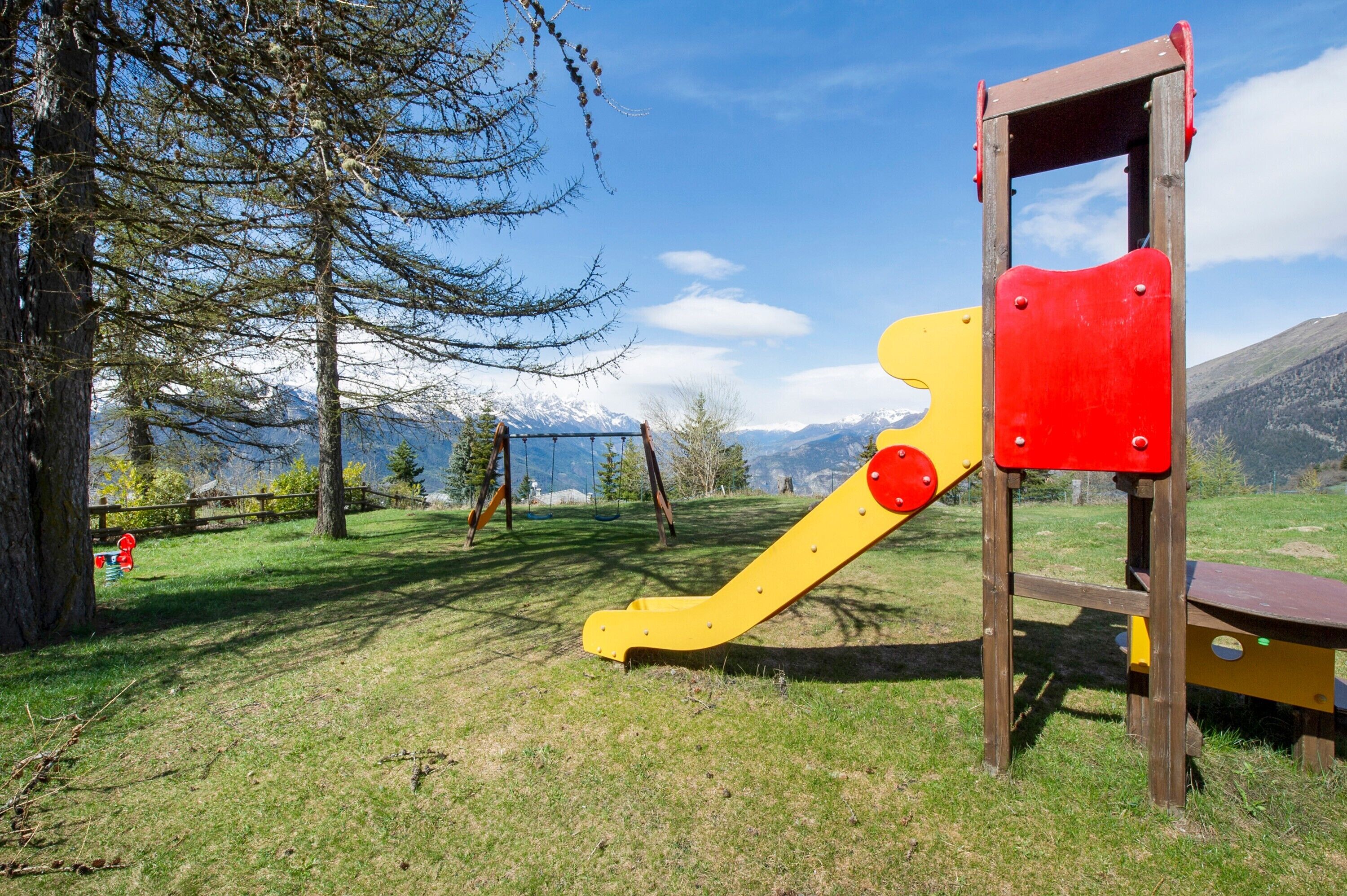 A playground with swings and a slide, surrounded by trees and mountains in the background.