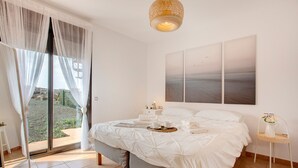 Un dormitorio con cama, mesita de noche y ventana grande con cortinas translúcidas, con vista al exterior.