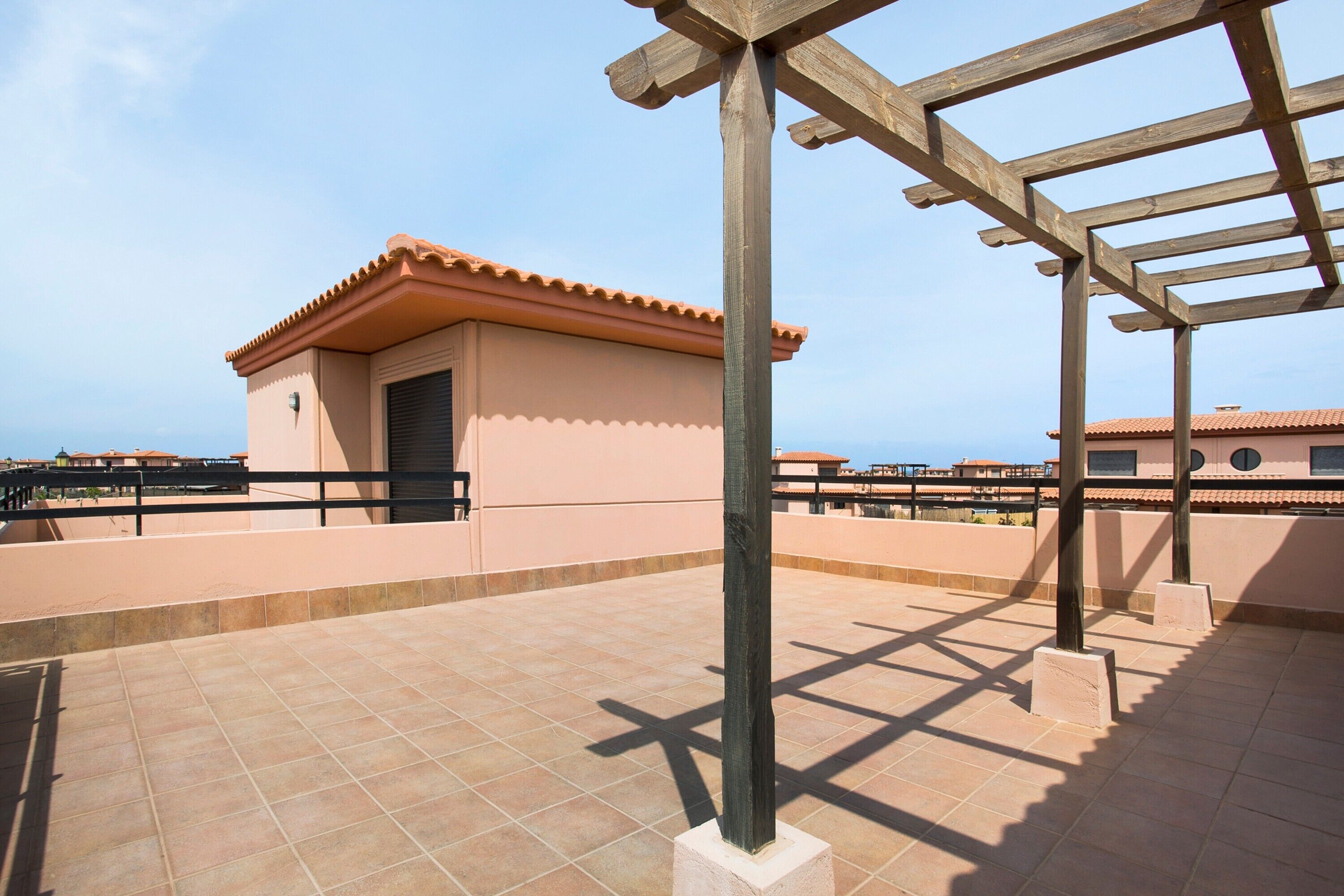 Una terraza en la azotea con una pérgola, un pequeño edificio de techo a tejas y un cielo despejado.