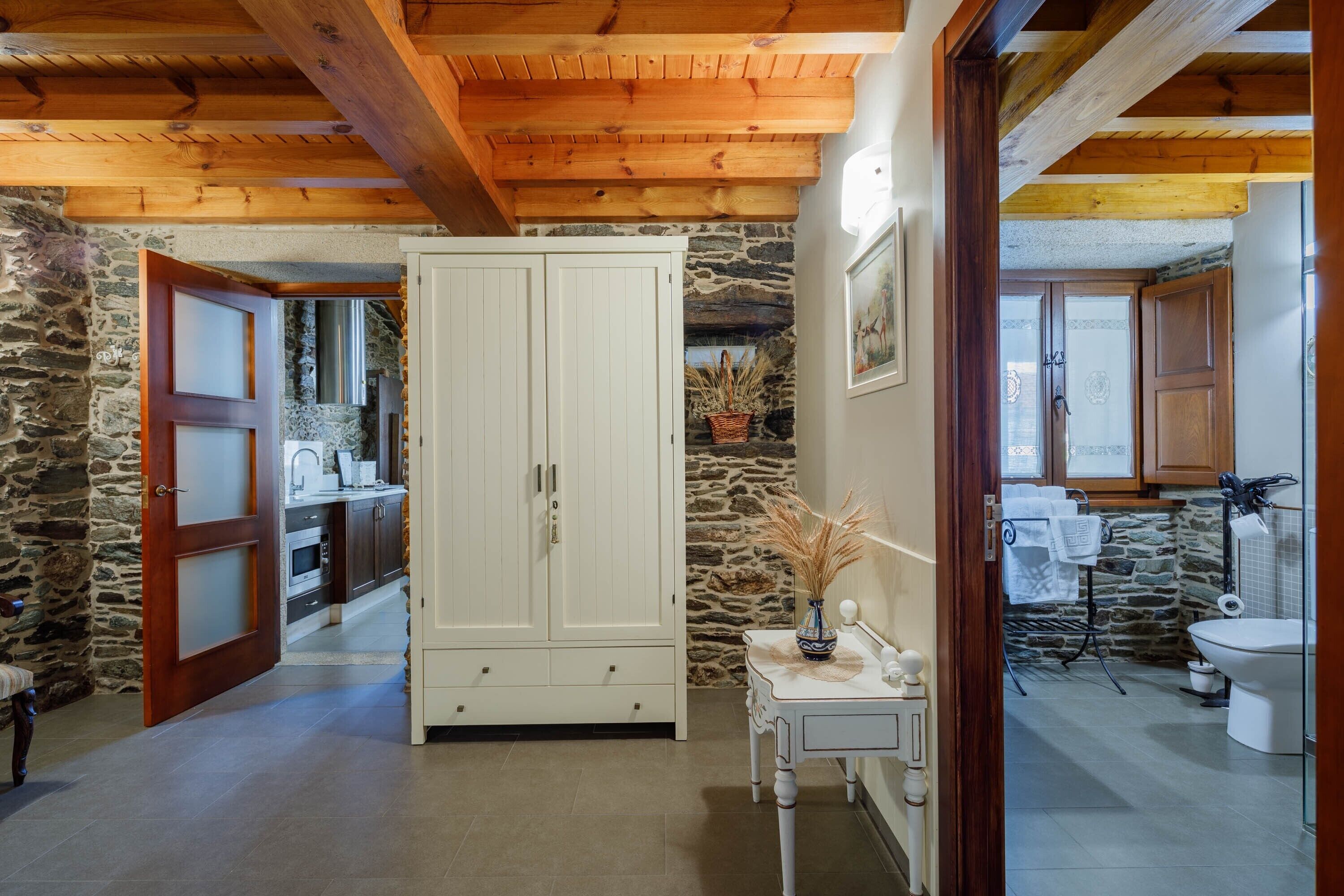 A rustic kitchen with stone walls, wooden beams, a white cabinet, and a small table with a vase.