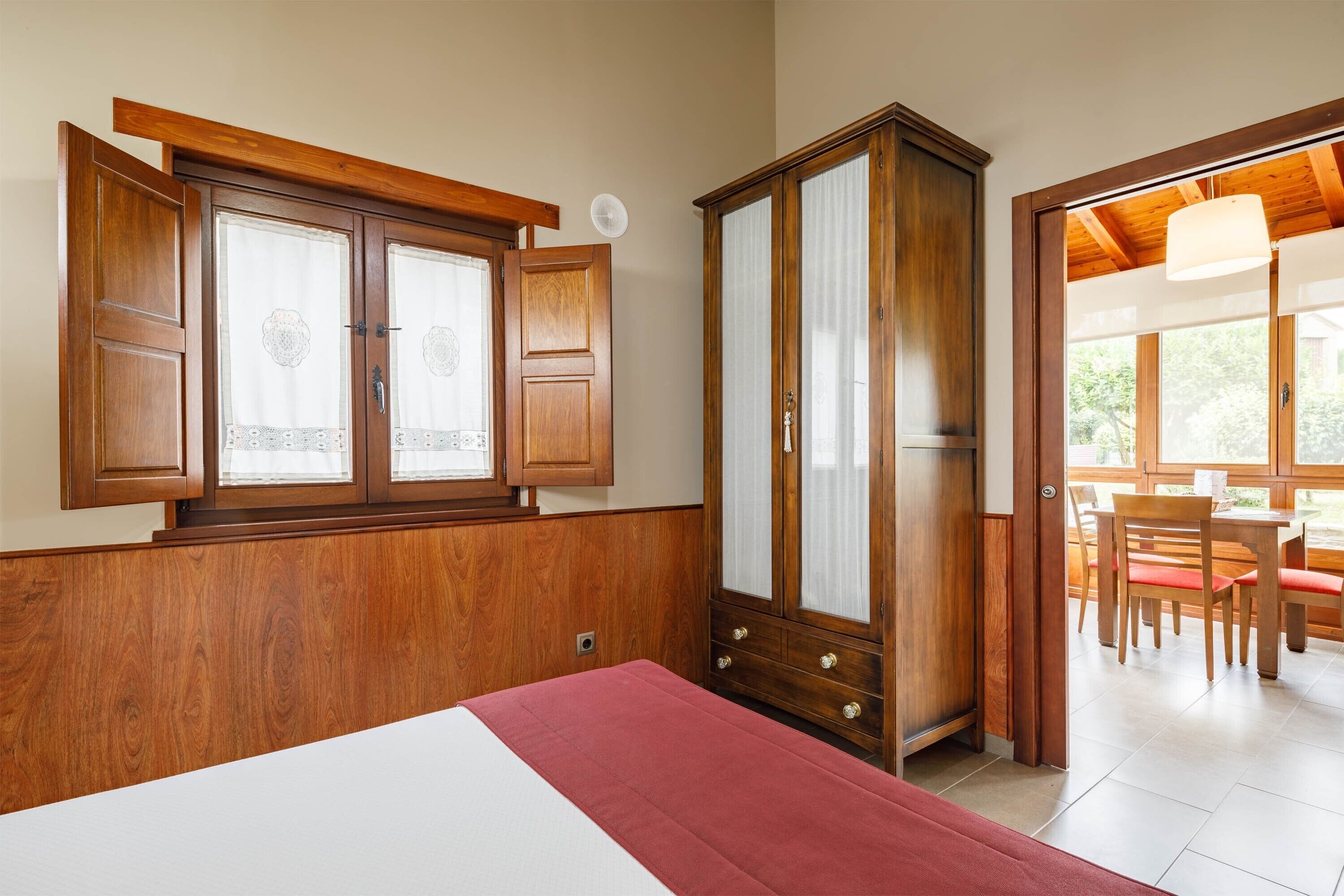 Um quarto com um guarda-roupa de madeira, uma cama com manta vermelha e uma janela com cortinas.