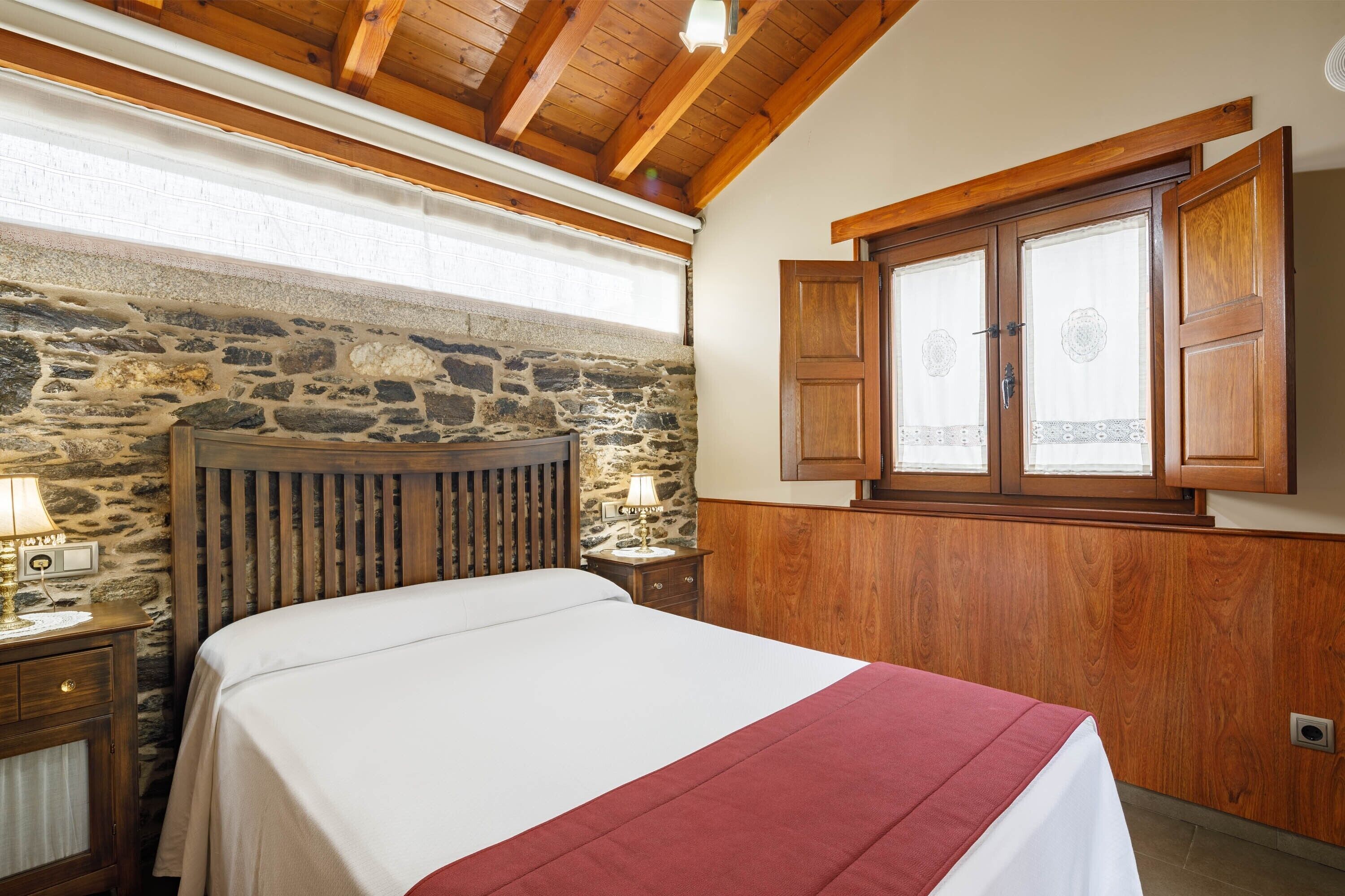 A bedroom with a stone wall, wooden ceiling, a bed with a white and red bedspread, a nightstand with a lamp, and a window with wooden shutters.
