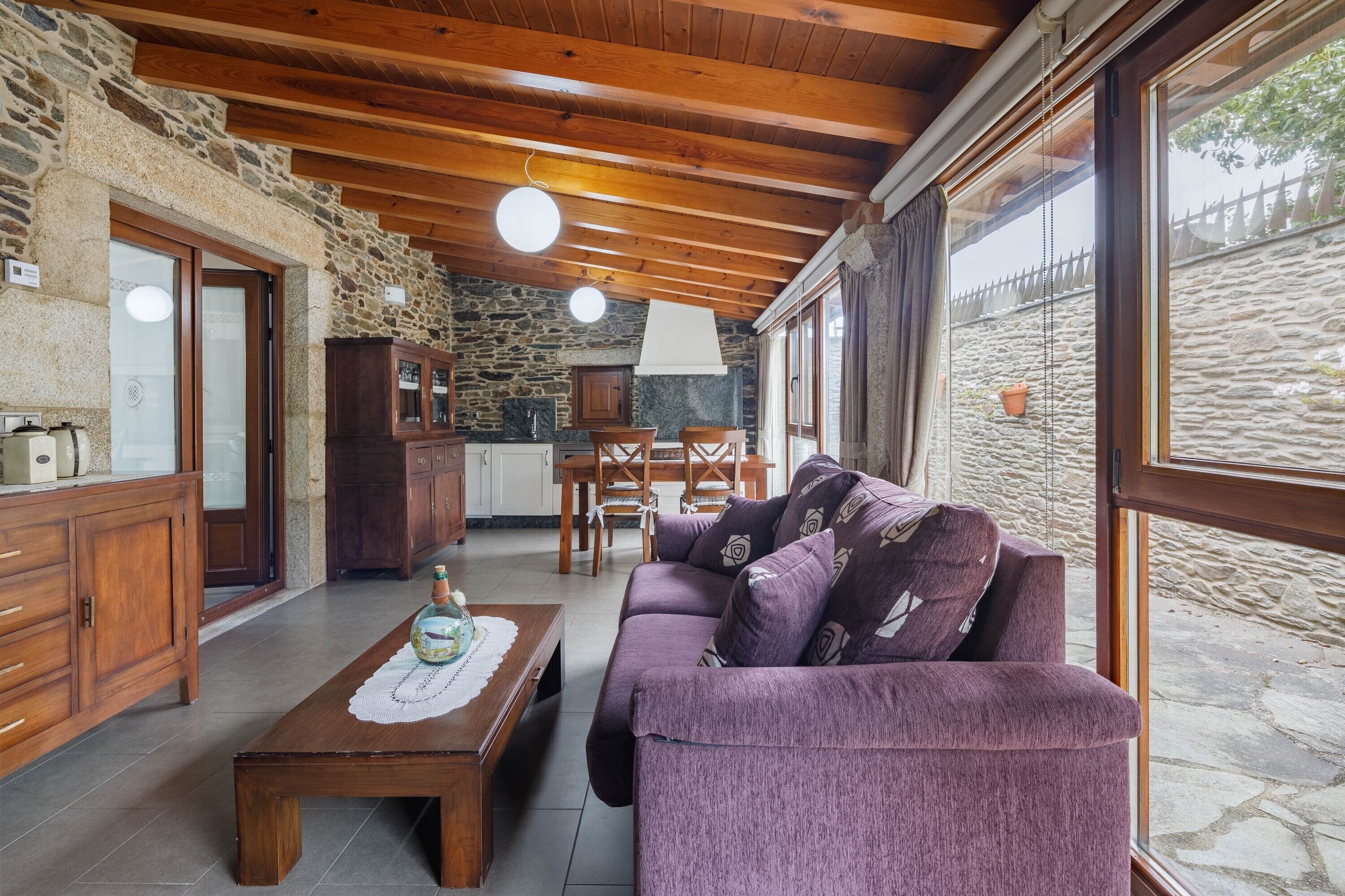 Un interior rústico con paredes de piedra, vigas de madera, un sofá morado, una mesa de centro de madera y una chimenea de piedra.