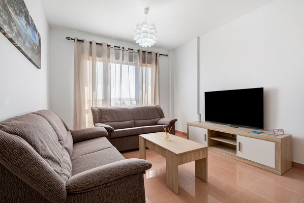 A living room with a sofa, a coffee table, a TV on a wooden stand, and a window with curtains.