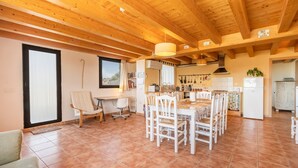 A spacious room with a wooden ceiling, a dining table with chairs, a sofa, and a kitchen area with a refrigerator and utensils. - Farmhouse 'El Refugi De Rubinat' with Mountain View, Wi-Fi and Air Conditioning (Rubinat)