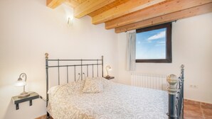 A bedroom with a bed, a nightstand with a lamp, a window with a view, and wooden ceiling beams. - Farmhouse 'El Refugi De Rubinat' with Mountain View, Wi-Fi and Air Conditioning (Rubinat)
