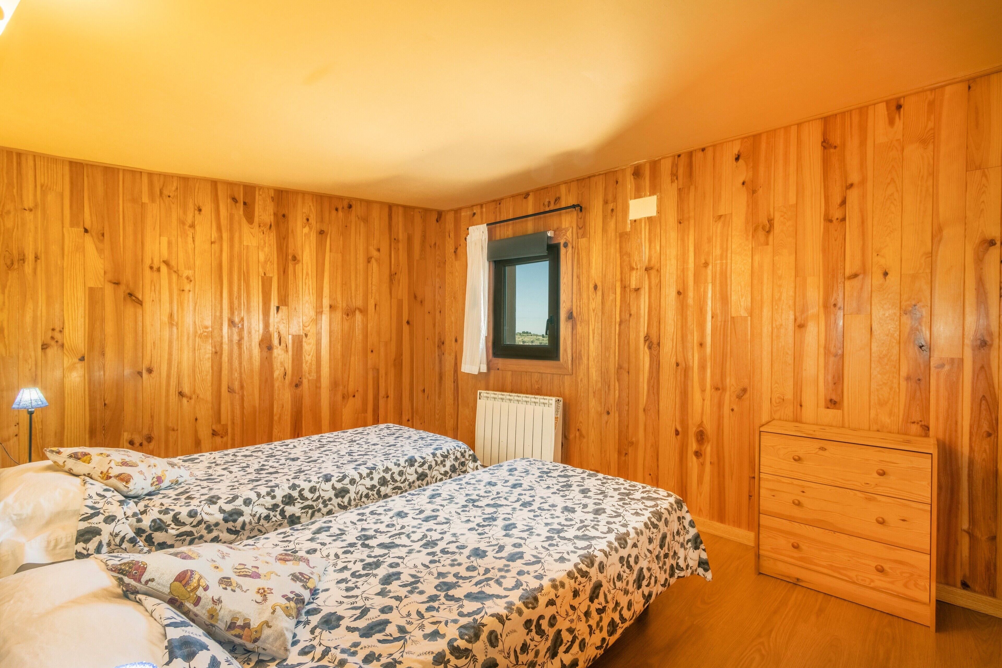 A room with wooden walls, two beds with floral bedding, a small window, a radiator, and a wooden dresser.