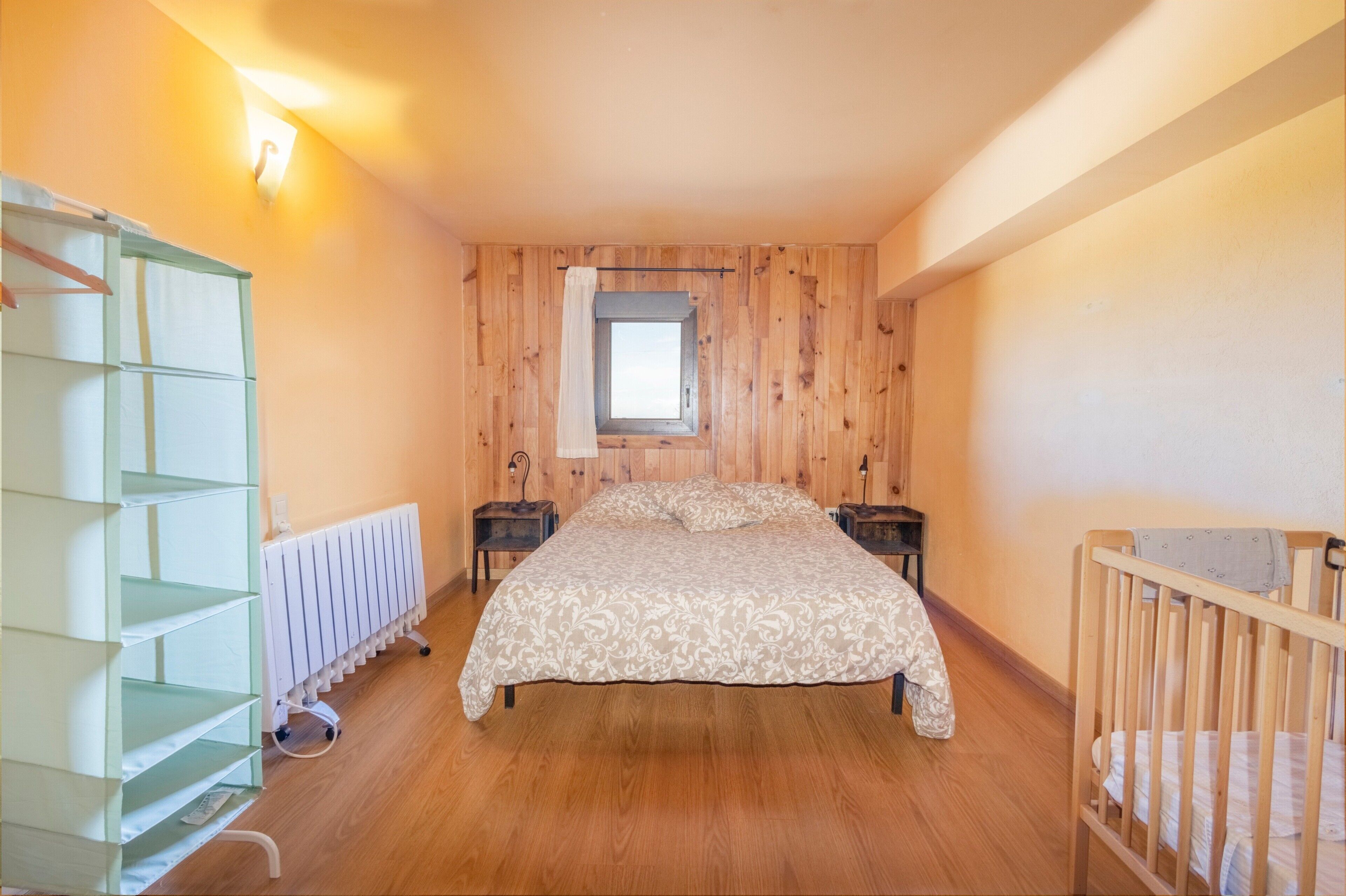 Un dormitorio con pared de paneles de madera, una ventana, una cama con colcha estampada, una mesita de noche, un radiador y una cuna.