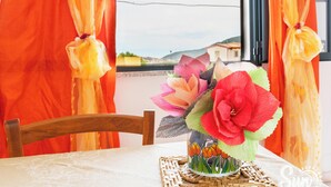 A table with a decorative vase, a wooden chair, and a view of a building through a window with orange curtains. - Holiday Home 'Limpiddu 4' with Mountain View, Shared Garden and Balcony (Limpiddu)