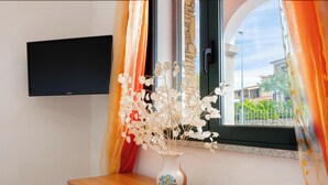Una stanza con una TV a schermo piatto montata a parete, un vaso con fiori bianchi su un tavolo e una finestra con tende.