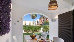 Une table de petit-déjeuner avec de la nourriture et des boissons, des chaises blanches, un luminaire suspendu en osier et une vue sur des bâtiments et de la verdure.