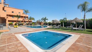 Un espace piscine comprenant deux piscines, des chaises longues, des palmiers et un bâtiment doté de balcons.