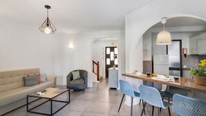 Un salon moderne comprenant un canapé, un fauteuil, une table basse et un coin repas avec des chaises, et une cuisine visible au fond.