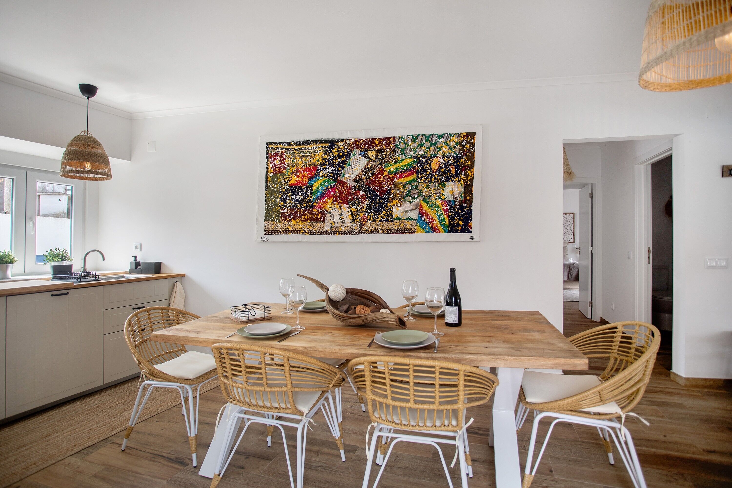 Zona pranzo con tavolo in legno, sedie in vimini, un grande quadro astratto e un lampadario.