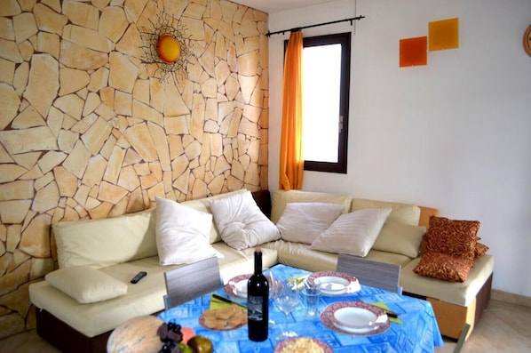 A living room with a stone wall, a sofa, a dining table with plates and a bottle, and a window with curtains.