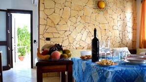 A dining area with a stone wall, a table set with a blue tablecloth, a bottle of wine, and a fruit bowl. - Villa 'Ll04 Hikari with Outdoor Spaces', Private Garden, Wi-Fi & Air Conditioning (San foca)