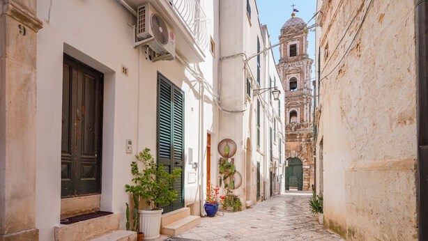 Une ruelle étroite pavée, bordée de bâtiments historiques, avec un clocher qui se profile au loin.