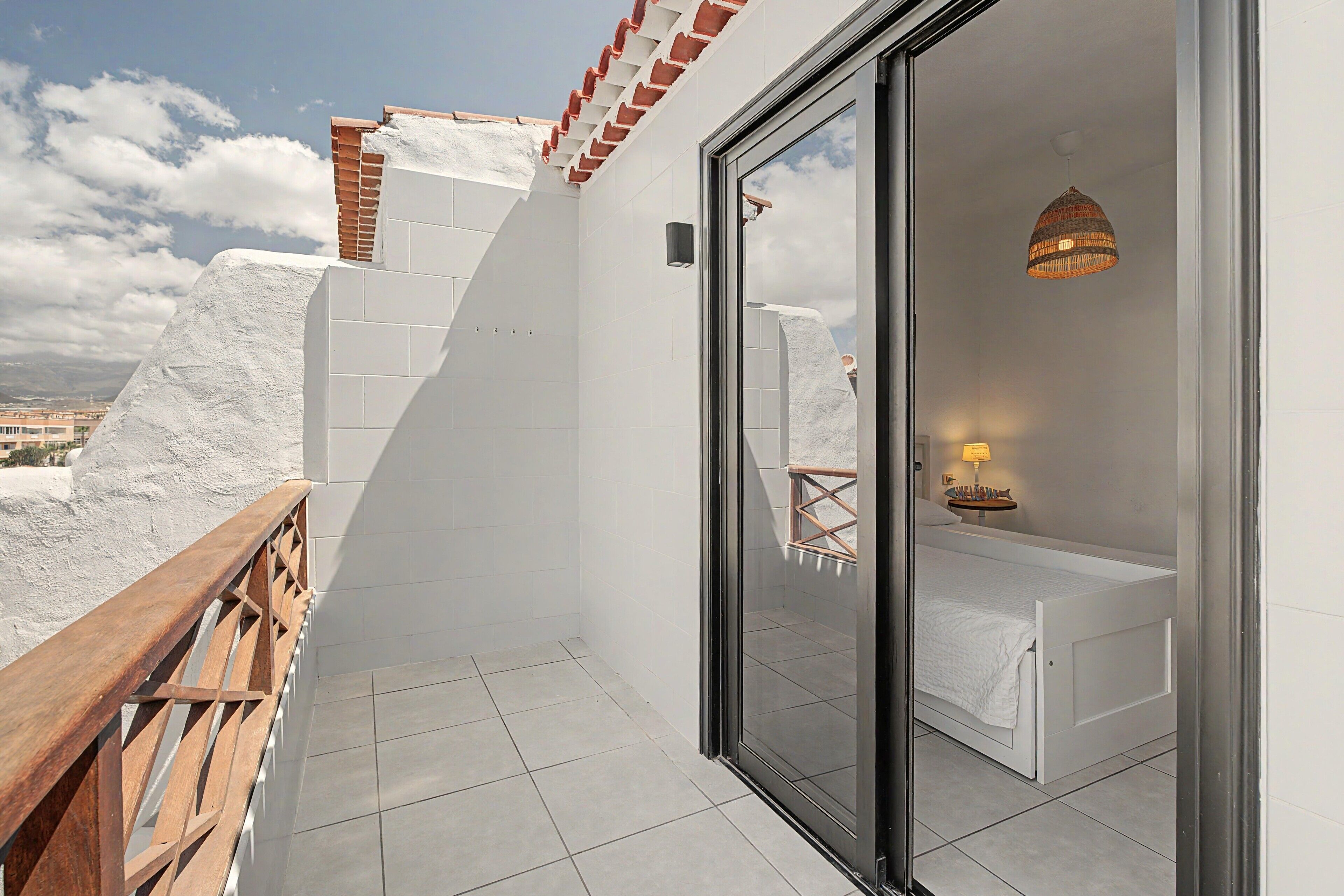 A balcony with a wooden railing, a white tiled floor, and a glass door leading to a room with a bed and a small table with a lamp.