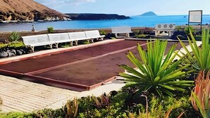 A beachside area with a long bench, a small garden with various plants, and a view of the sea and distant mountains. - Apartment with Stunning Ocean View, Pool, Garden, Terrace & Wi-Fi (Costa del Silencio)