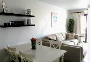 Una sala de estar con una mesa blanca, sillas, un sofá, una planta en maceta y un estante con recipientes.