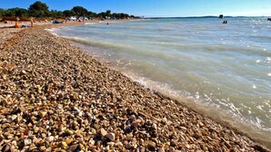Une plage de galets avec une eau claire, quelques personnes et des bateaux au loin.