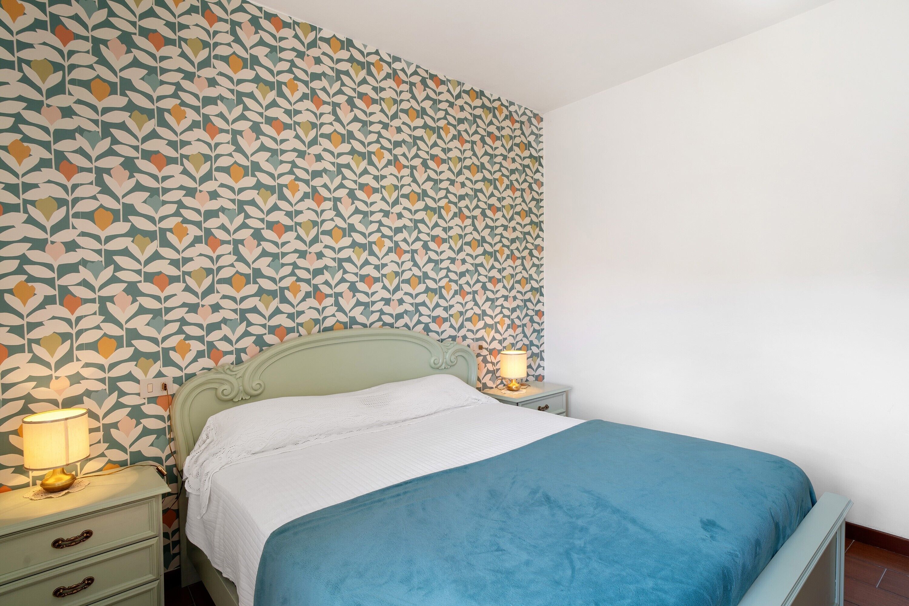 Quarto com papel de parede estampado, cama com colcha azul, mesa de cabeceira com abajur e cômoda com gavetas.