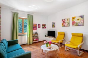 Un salon avec un canapé, deux fauteuils jaunes, un téléviseur et des œuvres d’art aux motifs floraux accrochées aux murs.