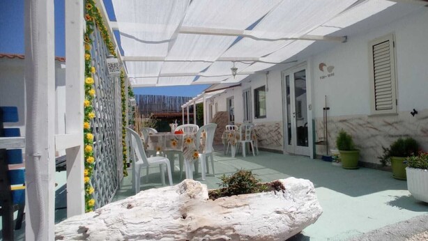 Área externa coberta com mesa, cadeiras, plantas em vasos e um objeto decorativo branco.