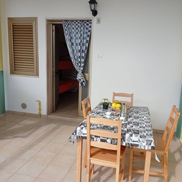 Un coin repas avec une table dressée pour deux personnes, un rideau à carreaux et une porte donnant sur une pièce où se trouve un lit.