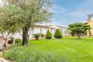 A well-maintained garden with various shrubs and a large tree, a white building with a tiled roof, and a clear sky.