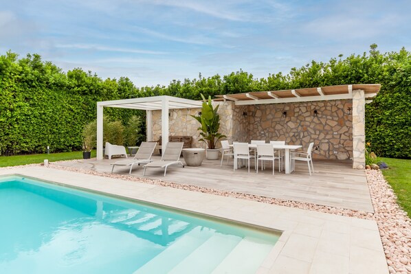 Un área junto a la piscina con una pérgola, un muro de piedra y mobiliario exterior.