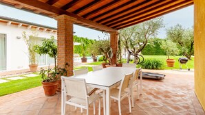 Une terrasse couverte avec une table et des chaises blanches, entourée de plantes en pot et offrant une vue sur un jardin.