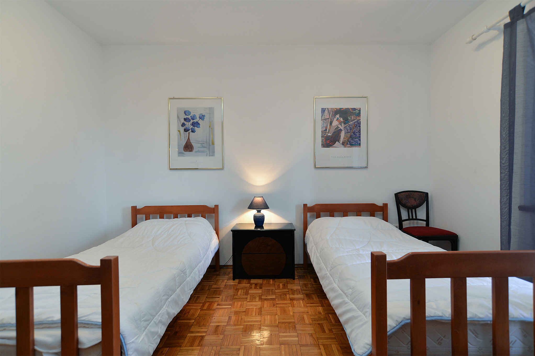 Una habitación con dos camas individuales, una mesita de noche con lámpara, una silla y dos cuadros en la pared.