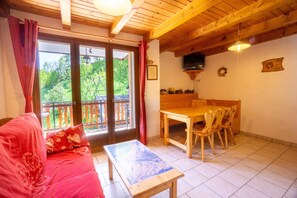 A cozy room with a wooden ceiling, a sofa with red cushions, a dining table with chairs, and a view of greenery outside.