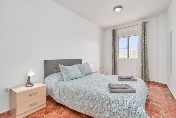 A bedroom with a bed, a bedside table with a lamp, a nightstand, and a window with curtains.