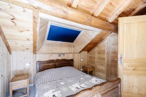 A wooden cabin bedroom with a sloped ceiling, a bed with a checkered blanket, a small wooden stool, and a wooden door.