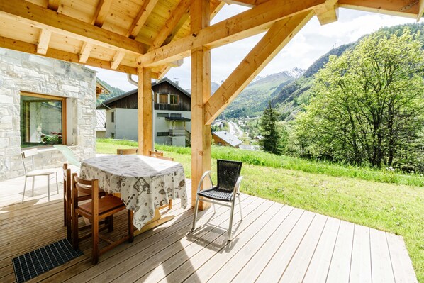 A wooden deck with a table and chairs, a stone wall, and a view of mountains and greenery.
